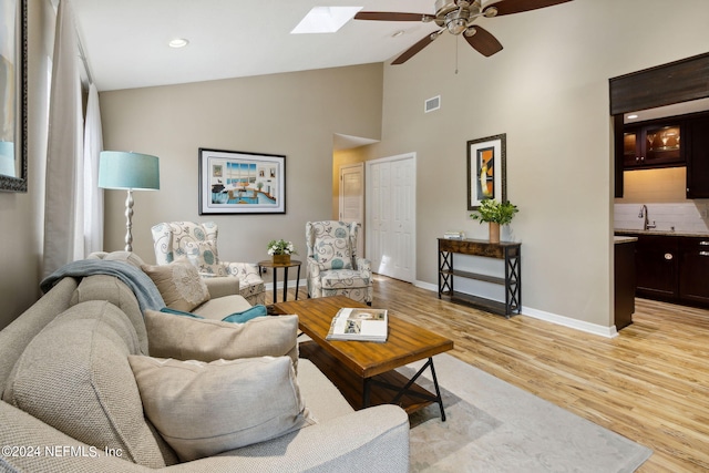 living room featuring high vaulted ceiling, ceiling fan, sink, light hardwood / wood-style floors, and a skylight