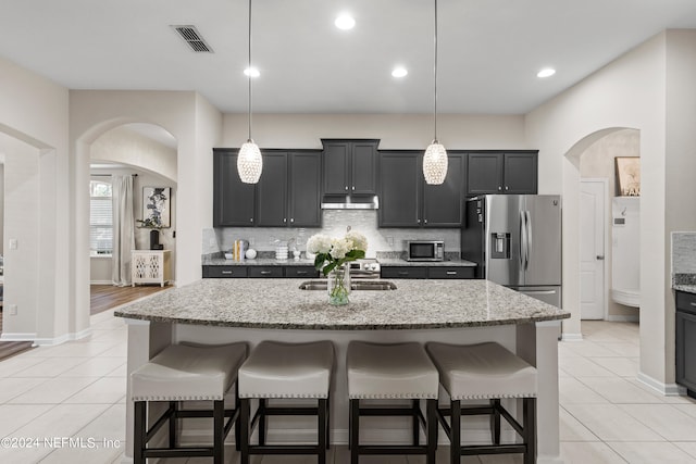 kitchen with light stone countertops, stainless steel refrigerator with ice dispenser, decorative light fixtures, and a kitchen island with sink