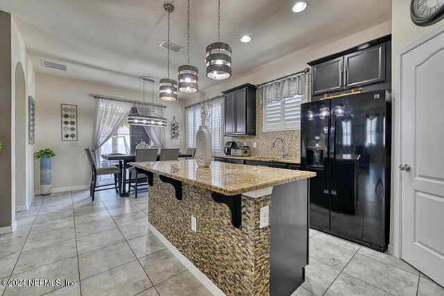 kitchen with a textured ceiling, an island with sink, hanging light fixtures, light stone countertops, and black refrigerator with ice dispenser