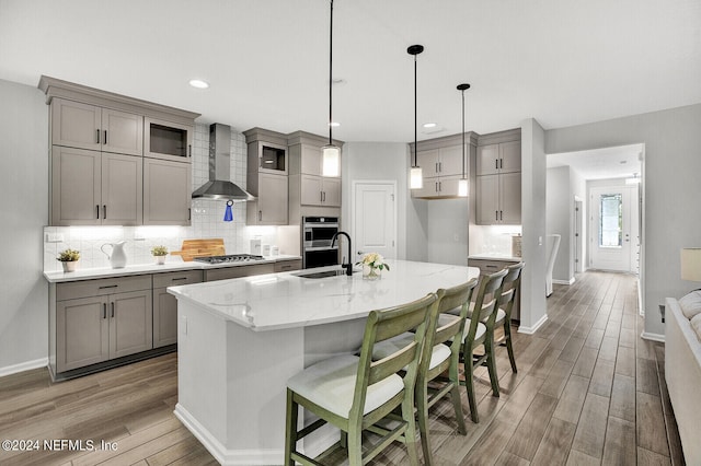 kitchen featuring wood-type flooring, a center island with sink, wall chimney exhaust hood, and light stone counters