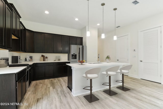 kitchen with an island with sink, hanging light fixtures, sink, stainless steel refrigerator with ice dispenser, and light wood-type flooring