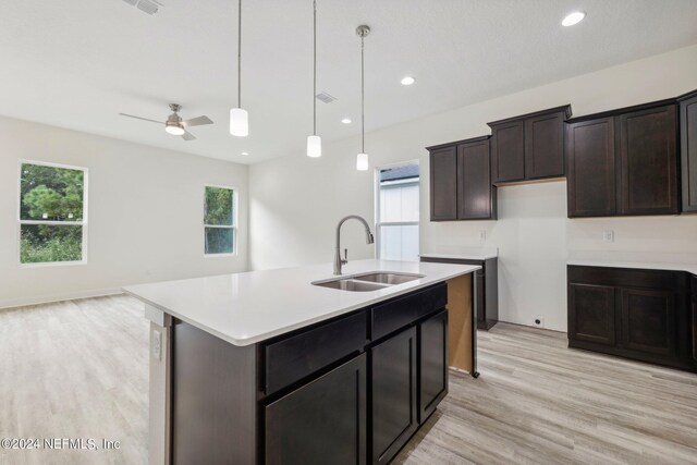 kitchen with hanging light fixtures, light wood-type flooring, dark brown cabinets, a kitchen island with sink, and sink