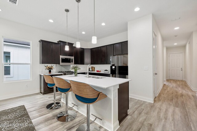 kitchen with an island with sink, sink, decorative light fixtures, appliances with stainless steel finishes, and light hardwood / wood-style floors