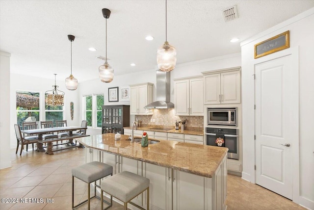 kitchen featuring appliances with stainless steel finishes, a kitchen island with sink, decorative light fixtures, and wall chimney range hood