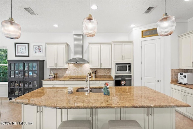 kitchen with wall chimney range hood, a large island with sink, appliances with stainless steel finishes, a textured ceiling, and sink