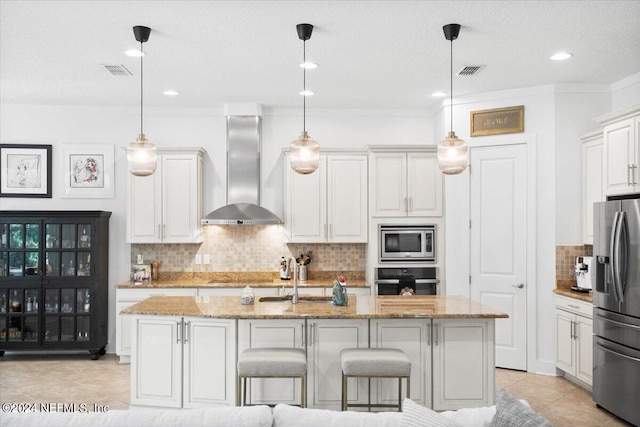 kitchen featuring stainless steel appliances, wall chimney exhaust hood, an island with sink, and hanging light fixtures