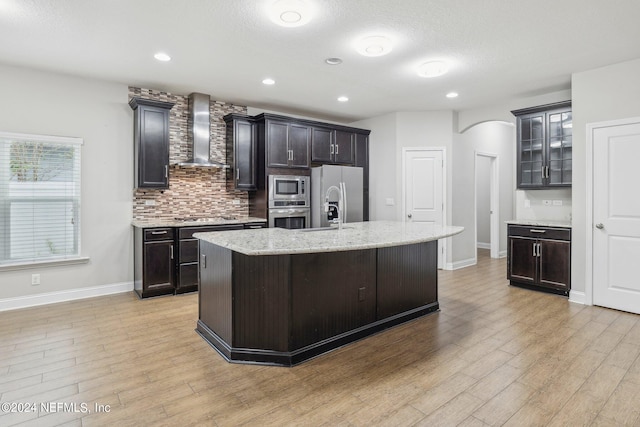 kitchen featuring light wood-type flooring, wall chimney exhaust hood, stainless steel appliances, a center island with sink, and light stone countertops