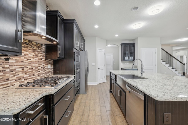 kitchen with sink, a center island with sink, wall chimney exhaust hood, appliances with stainless steel finishes, and light wood-type flooring