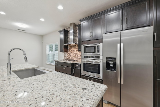 kitchen with sink, wall chimney exhaust hood, stainless steel appliances, backsplash, and light stone countertops