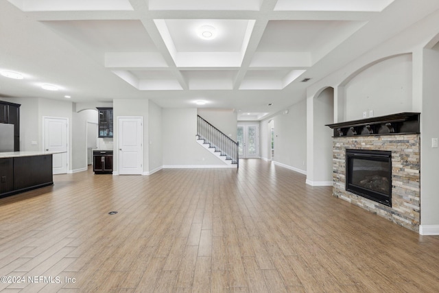 unfurnished living room featuring light hardwood / wood-style flooring, beamed ceiling, coffered ceiling, and a stone fireplace