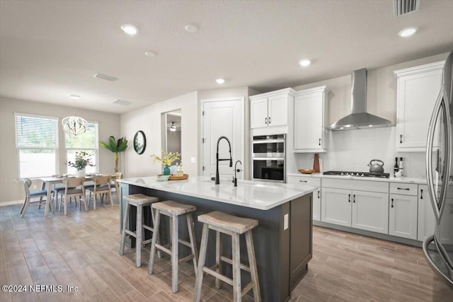 kitchen with wall chimney range hood, appliances with stainless steel finishes, hanging light fixtures, light hardwood / wood-style flooring, and a center island with sink