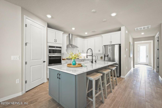 kitchen featuring wall chimney range hood, white cabinetry, a kitchen island with sink, a kitchen bar, and stainless steel appliances