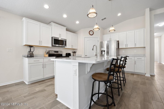 kitchen with an island with sink, hanging light fixtures, white cabinetry, appliances with stainless steel finishes, and vaulted ceiling