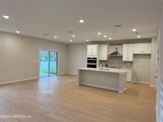 kitchen with light wood-type flooring, wall chimney exhaust hood, a kitchen island with sink, sink, and white cabinets