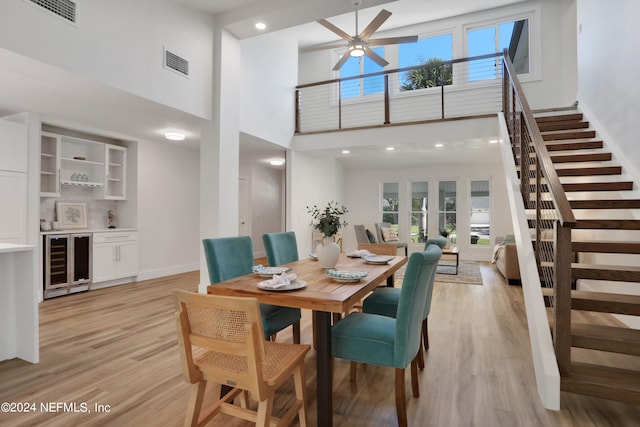 dining room with beverage cooler, ceiling fan, indoor bar, light wood-type flooring, and a high ceiling