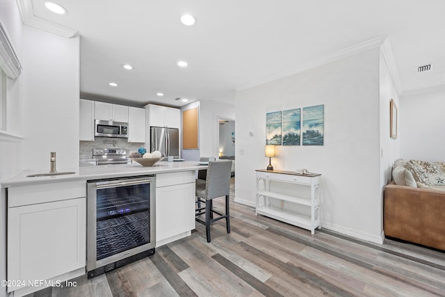 kitchen with appliances with stainless steel finishes, wine cooler, white cabinets, crown molding, and hardwood / wood-style flooring