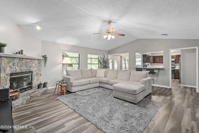 living room with wood-type flooring, a textured ceiling, vaulted ceiling, a stone fireplace, and ceiling fan