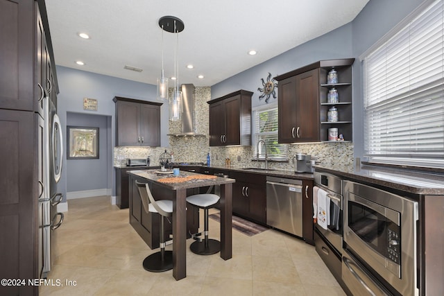 kitchen featuring pendant lighting, a kitchen island, wall chimney range hood, appliances with stainless steel finishes, and dark brown cabinetry