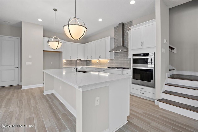 kitchen featuring white cabinets, sink, a center island with sink, wall chimney range hood, and appliances with stainless steel finishes