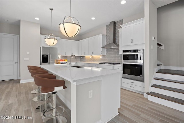 kitchen with sink, an island with sink, white cabinets, wall chimney exhaust hood, and appliances with stainless steel finishes