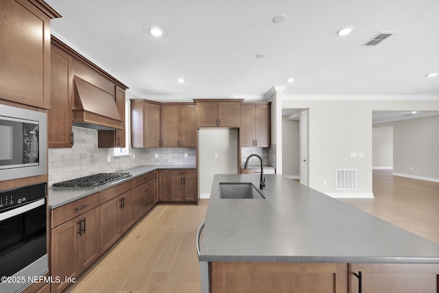 kitchen featuring light wood-type flooring, custom range hood, stainless steel appliances, sink, and an island with sink