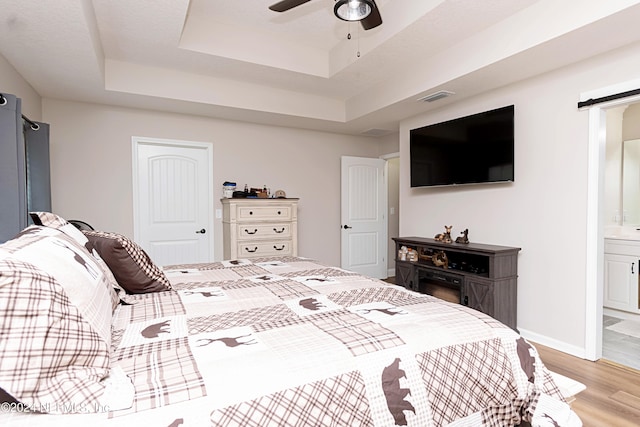 bedroom with ceiling fan, a barn door, a tray ceiling, light hardwood / wood-style flooring, and ensuite bath