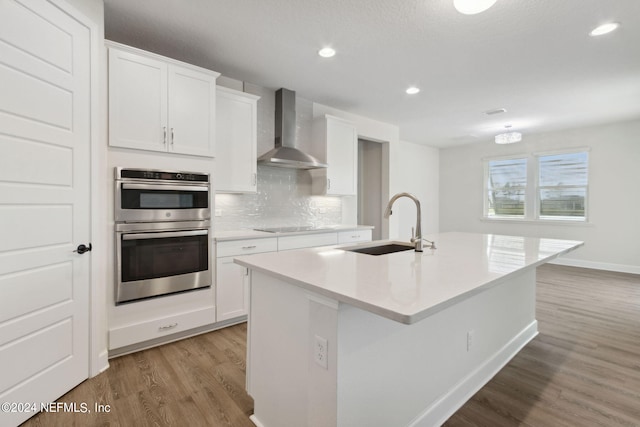 kitchen with light wood-type flooring, wall chimney exhaust hood, stainless steel double oven, and a sink