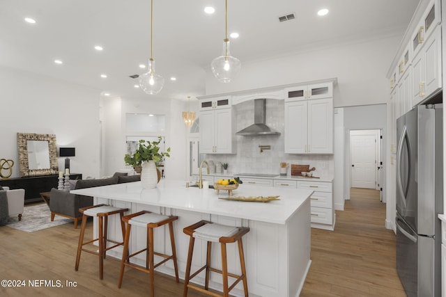 kitchen with a large island with sink, wall chimney range hood, light hardwood / wood-style flooring, white cabinetry, and stainless steel refrigerator