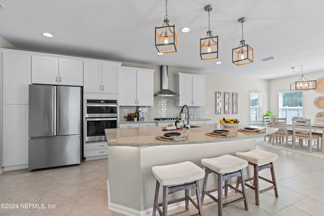 kitchen featuring a kitchen island with sink, stainless steel appliances, wall chimney exhaust hood, white cabinetry, and backsplash