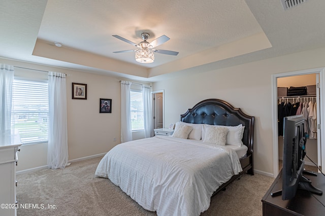 carpeted bedroom with a tray ceiling, a walk in closet, multiple windows, and ceiling fan