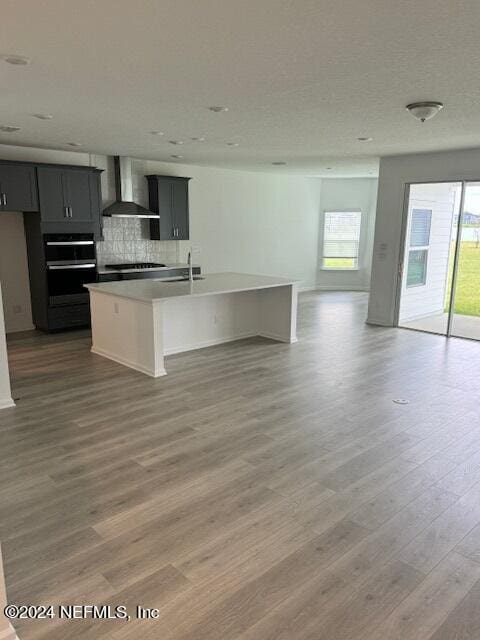 kitchen with decorative backsplash, wood-type flooring, wall chimney exhaust hood, sink, and a center island with sink