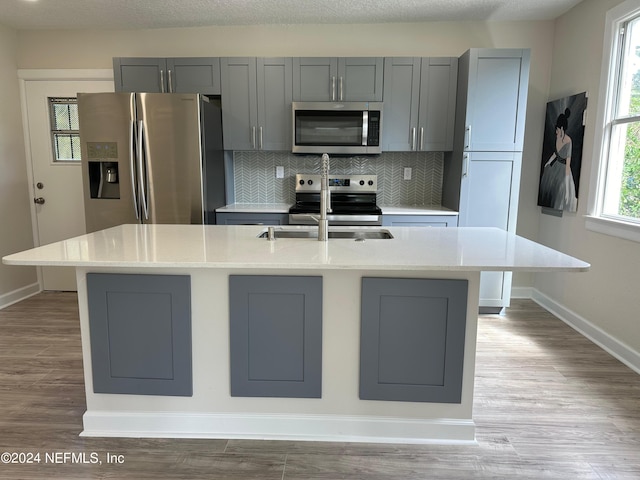 kitchen featuring appliances with stainless steel finishes, light wood-type flooring, gray cabinets, and a kitchen island with sink