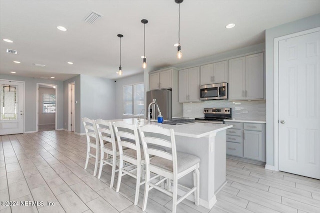 kitchen featuring gray cabinetry, decorative light fixtures, a center island with sink, backsplash, and appliances with stainless steel finishes