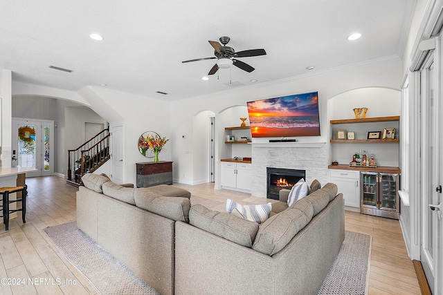 living room with light wood-type flooring, wine cooler, a stone fireplace, and crown molding