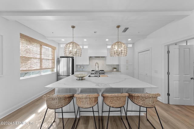 kitchen with light stone counters, white cabinets, stainless steel fridge, a center island with sink, and decorative light fixtures