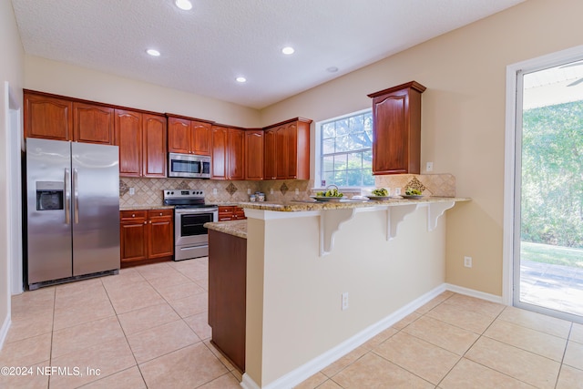 kitchen with kitchen peninsula, appliances with stainless steel finishes, a kitchen breakfast bar, light tile patterned flooring, and light stone counters