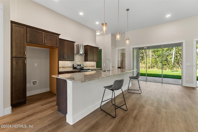 kitchen with wall chimney range hood, a center island with sink, stainless steel range with electric stovetop, light stone counters, and light hardwood / wood-style floors