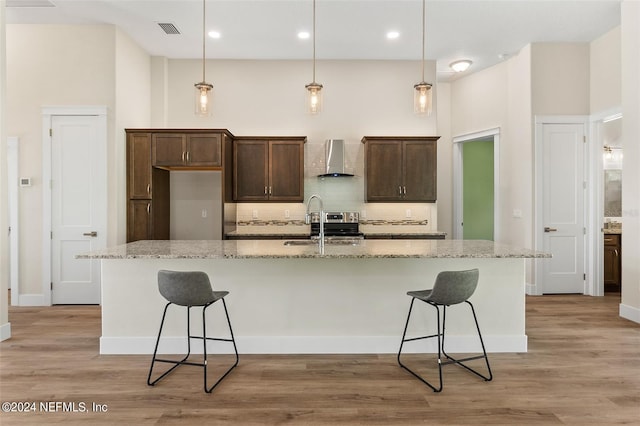 kitchen featuring wall chimney exhaust hood, light hardwood / wood-style flooring, an island with sink, pendant lighting, and light stone counters