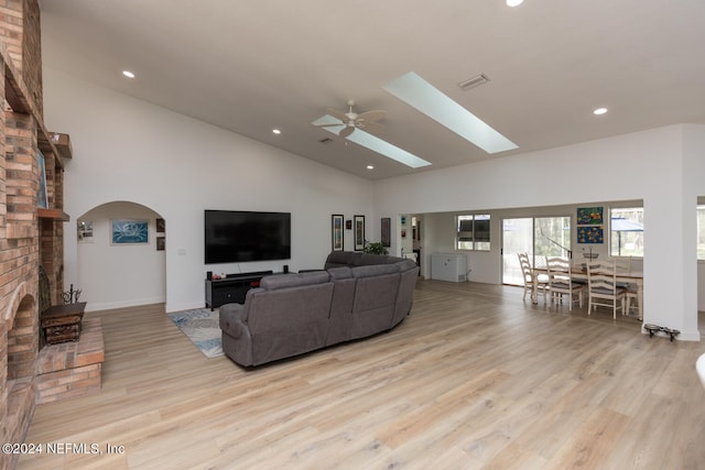 living room featuring a skylight, a brick fireplace, light hardwood / wood-style floors, ceiling fan, and high vaulted ceiling