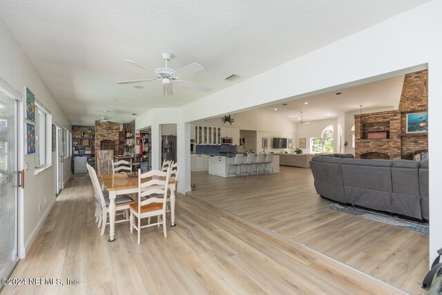 dining space featuring a brick fireplace, a textured ceiling, light hardwood / wood-style floors, and ceiling fan