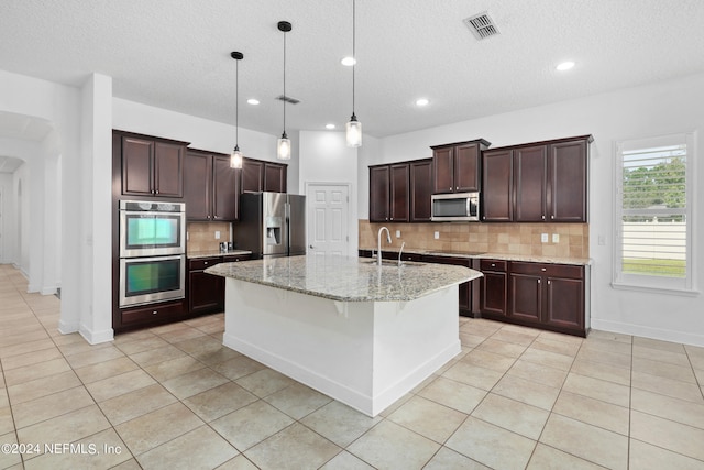 kitchen featuring light stone counters, an island with sink, pendant lighting, decorative backsplash, and appliances with stainless steel finishes