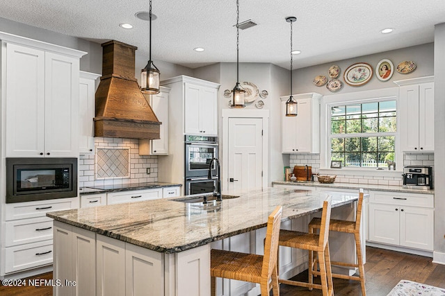 kitchen with custom range hood, a textured ceiling, decorative light fixtures, and a center island with sink