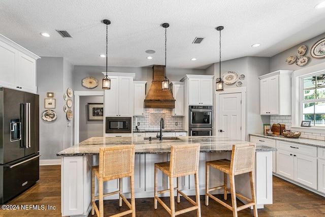 kitchen with dark hardwood / wood-style floors, custom range hood, a center island with sink, decorative light fixtures, and appliances with stainless steel finishes