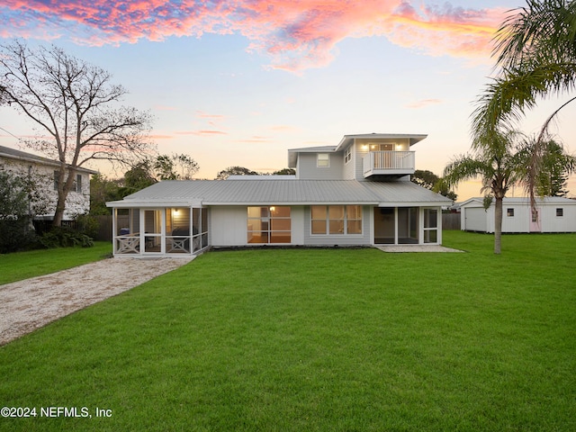 back house at dusk with a balcony, a lawn, and a sunroom