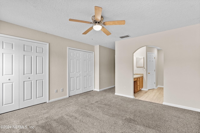 unfurnished bedroom with light carpet, two closets, ceiling fan, a textured ceiling, and ensuite bathroom