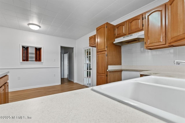 kitchen featuring backsplash, brown cabinets, wood finished floors, light countertops, and under cabinet range hood