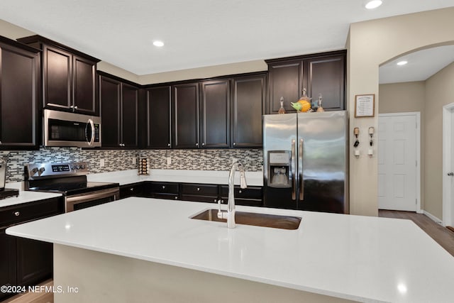 kitchen with dark brown cabinets, backsplash, appliances with stainless steel finishes, a kitchen island with sink, and sink