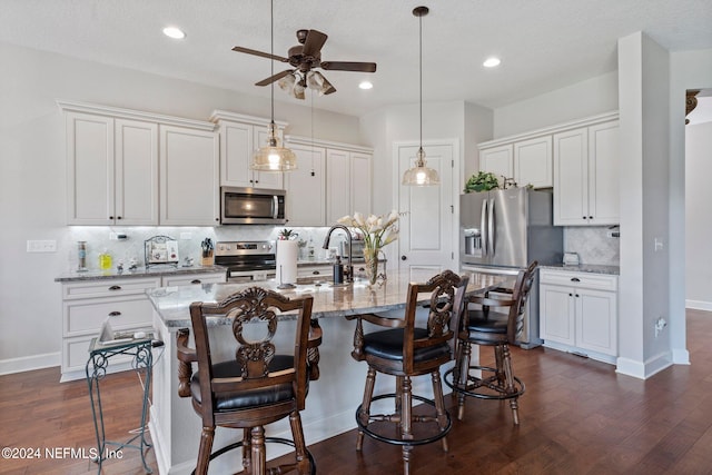 kitchen with light stone countertops, dark hardwood / wood-style flooring, stainless steel appliances, white cabinets, and a center island with sink