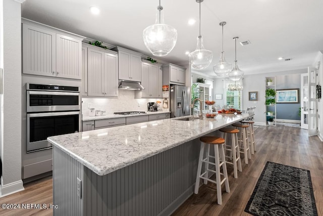 kitchen with a large island with sink, gray cabinetry, pendant lighting, appliances with stainless steel finishes, and dark hardwood / wood-style flooring