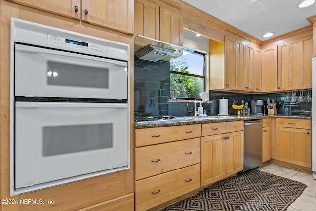 kitchen with decorative backsplash, stainless steel gas cooktop, dark stone counters, light tile patterned flooring, and white double oven